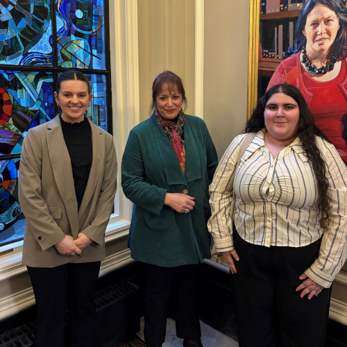 Two students and their supervisor pose flanked by a stain glass window and a large portrait painting. Two students and their supervisor pose flanked by a stain glass window and a large portrait painting.