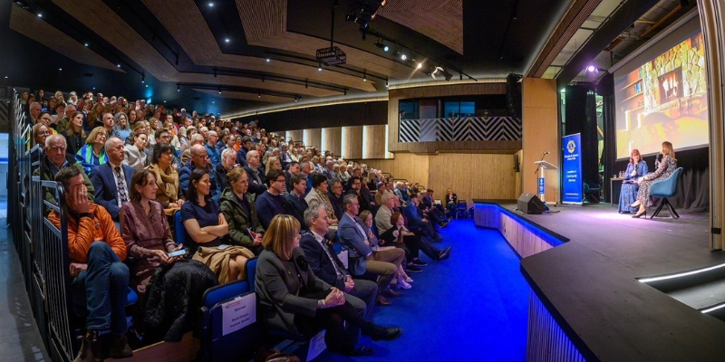 Side shot of two women sitting down on stage, in front of a packed theatre audience Side shot of two women sitting down on stage, in front of a packed theatre audience