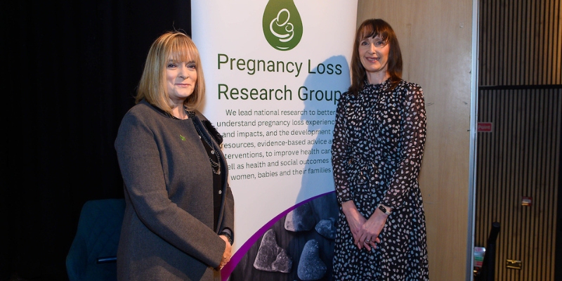 Two women standing smiling in between a pull up banner with 'Pregnancy Loss Research Group' written on it & the Group logo Two women standing smiling in between a pull up banner with 'Pregnancy Loss Research Group' written on it & the Group logo