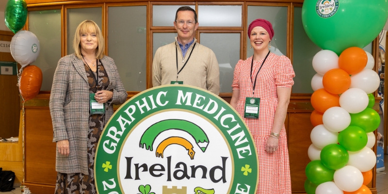 Three people standing behind a large circular sign for the Graphic Medicine Conference 2024, Ireland