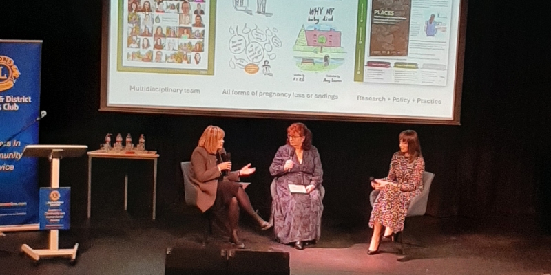 Three women sitting on stage having a fireside conversation. In the background is a large presentation screen displaying images and outputs from the Pregnancy Loss Research Group Three women sitting on stage having a fireside conversation. In the background is a large presentation screen displaying images and outputs from the Pregnancy Loss Research Group