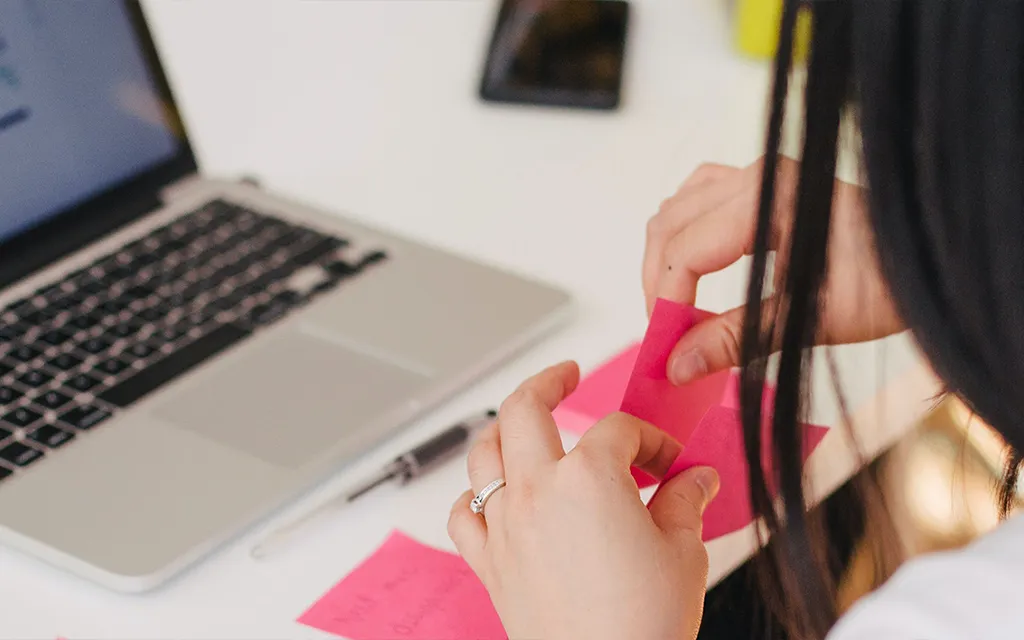 A female student holding pink post-its with a laptop on a desk.