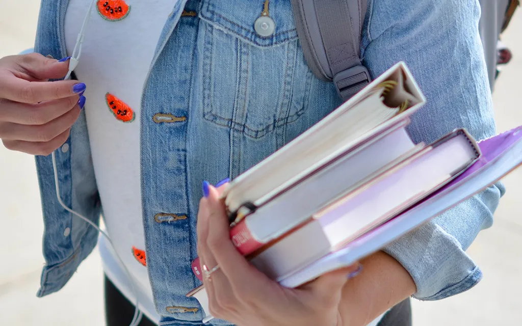 A student wearing a denim jacket and holding books and a folder.