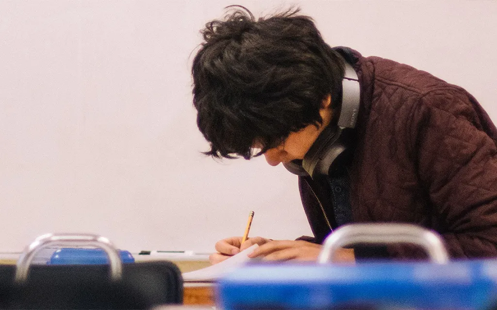 A student wearing a brown jacket sitting in a classroom writing.